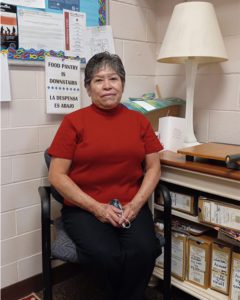 A middle aged Hispanic woman sits in a side chair in an office with cinder block walls. She has greying short dark hair with bangs and wears a red short sleeved shirt and black pants.