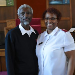 A Black couple smile at the camera inside a chapel. The man, on left, has white short hair and a goatee. He wears a white t-shirt, button down white shirt, and navy blue sweater. The woman, on right, has short black hair and glasses. She wears a white button down blouse with red officer epaulets.