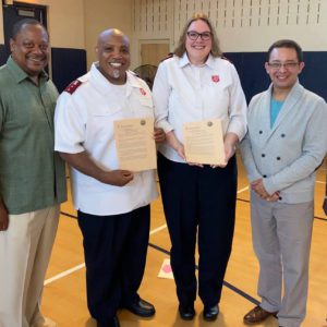 Four smiling adults standing on an indoor basketball court. Left to right: an African American man with short hair, olive green button down shirt, and tan trousers; a bald African American man with goatee, white Salvation Army officer shirt, black trousers, holding a certificate; a tall white woman with shoulder length blonde hair, dressed like her husband to the left, also holding a certificate; and a Hispanic man with short dark hair, glasses, blue t-shirt, grey double breasted cardigan sweater, and trousers.