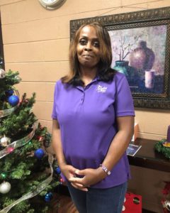 A smiling African American woman with long shoulder length hair wearing a purple polo shirt. She stands next to a decorated Christmas tree and a framed still life print can be seen behind her hanging from a peach color painted cinder block wall.