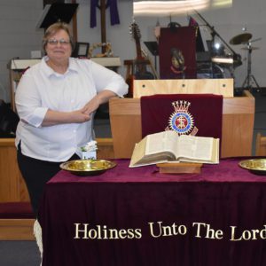 A woman wearing a white button down blouse and black pants stands next to a Christian altar. A Bible and holiness cloth, with embroidered text "Holiness Unto the Lord" and Salvation Army crest are visible on the altar.