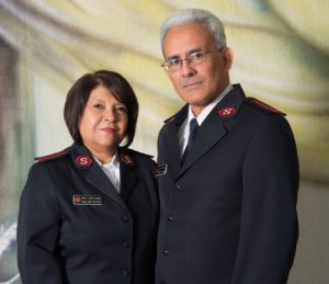 A Hispanic couple wearing Salvation Army uniforms consisting of a white button down shirt and navy suit jacket standing in front of a fabric studio background. The woman, on left, has short dark hair. The man, on right, has white hair and glasses.