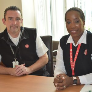 A smiling couple seated at a wood office table. The man on left is White with short dark hair and a salt-and-pepper goatee. He wears a white short sleeved shirt and zip up navy fleece vest with Red Shield logo embroidered on the chest. The woman, on right, is dressed similarly to her husband on left. She has dark hair which is pulled back from her face. She also wears a red Salvation Army lanyard around her neck.