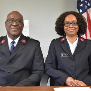 A smiling African American couple, both seated in front of a table with United States flag visible in background. The man, on left, is middle aged, bald, wears glasses, and is dressed in a Salvation Army officer uniform. The woman, on right, is also middle aged, has curly chin length dark hair, wears glasses, and is also dressed in a Salvation Army officer uniform.