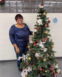 A middle aged African American woman with short dark hair, large hoop earrings, and glasses, wearing a blue shirt and pants poses with a decorated Christmas tree