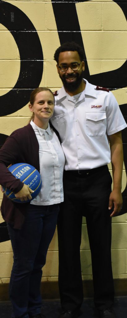 A White woman and African American man stand together in front of a painted wall. The woman holds a blue basketball. She wears a white button down shirt, black cardigan sweater, and black pants. Her long dark blonde hair is pulled into a low ponytail. The man has short hair and wears a while button down shirt with red Salvation Army officer epaulets and black pants.