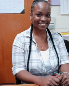 A smiling African American woman with hair pulled back and two long braids laying over her shoulders. She wears a white tank top and elbow length pastel plaid button down shirt. She is seated at a desk in an office.