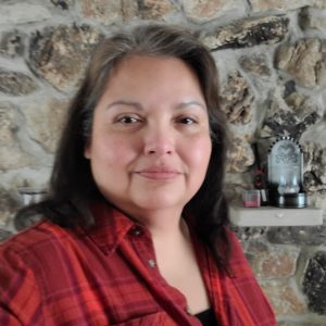 A Native American woman with a touch of grey in her hair wears a red plaid flannel shirt and stands in front of a rock fireplace or wall.
