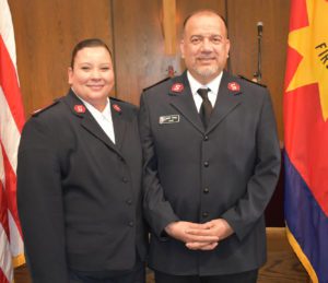 Photograph of a Hispanic couple. The woman, on left, has her long dark hair pulled back and wears a Salvation Army officer uniform. The man, on right, also wears the uniform. The couple are smiling at the camera and pose in front of a wood paneled wall with United States flag to the left and Salvation Army flag on right.