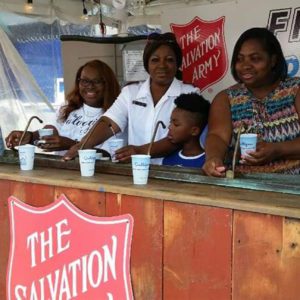 Three African American women and one African American boy stand behind a rustic wood serving counter. A Red Shield logo is attached to the front of the counter and can be seen on a white banner behind the counter. The woman second from left wears a Salvation Army officer uniform. The others wear casual clothing.