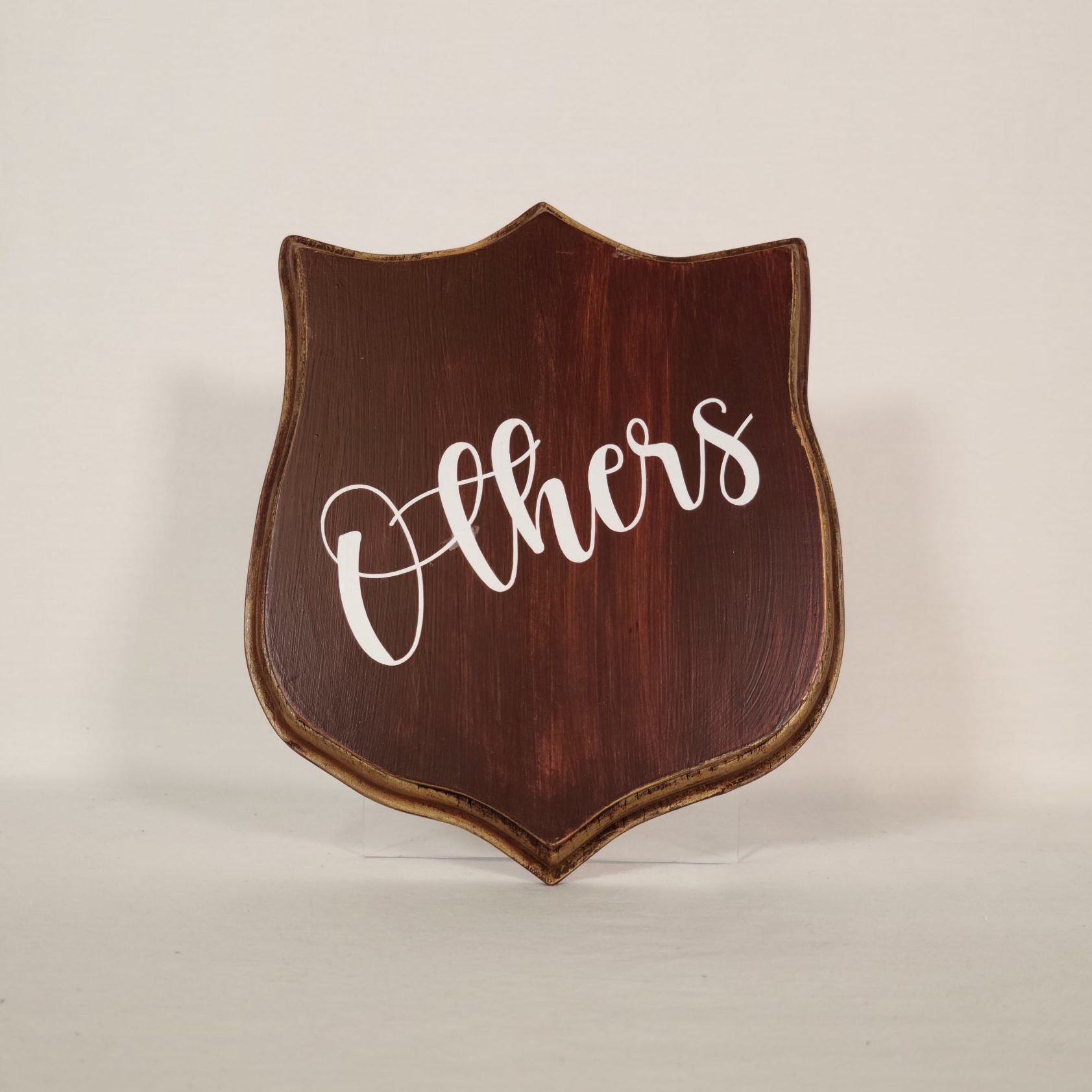 A brown wood shield shaped plaque with the word "others" written on it in white cursive font.