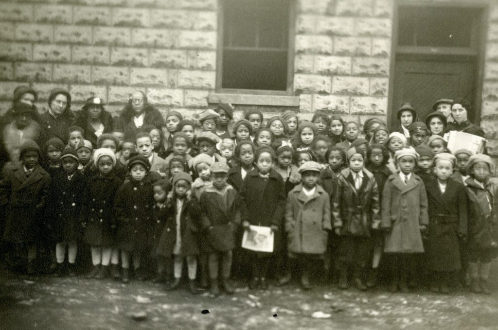 A black and white photograph showing a large group of African American children and adults along with some White female Salvationists, all posing in front of a stone building.