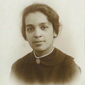A black and white photograph showing a light skinned mixed race woman with her hair pulled back into a bun who is wearing a Salvation Army uniform with shield shaped pin at the neck.
