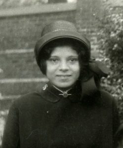 A black and white photo of a Hispanic woman shyly smiling at the camera. She wears a Salvation Army bonnet with the bow tied at her left ear, a Salvation Army uniform, and a bar pin at her neck.