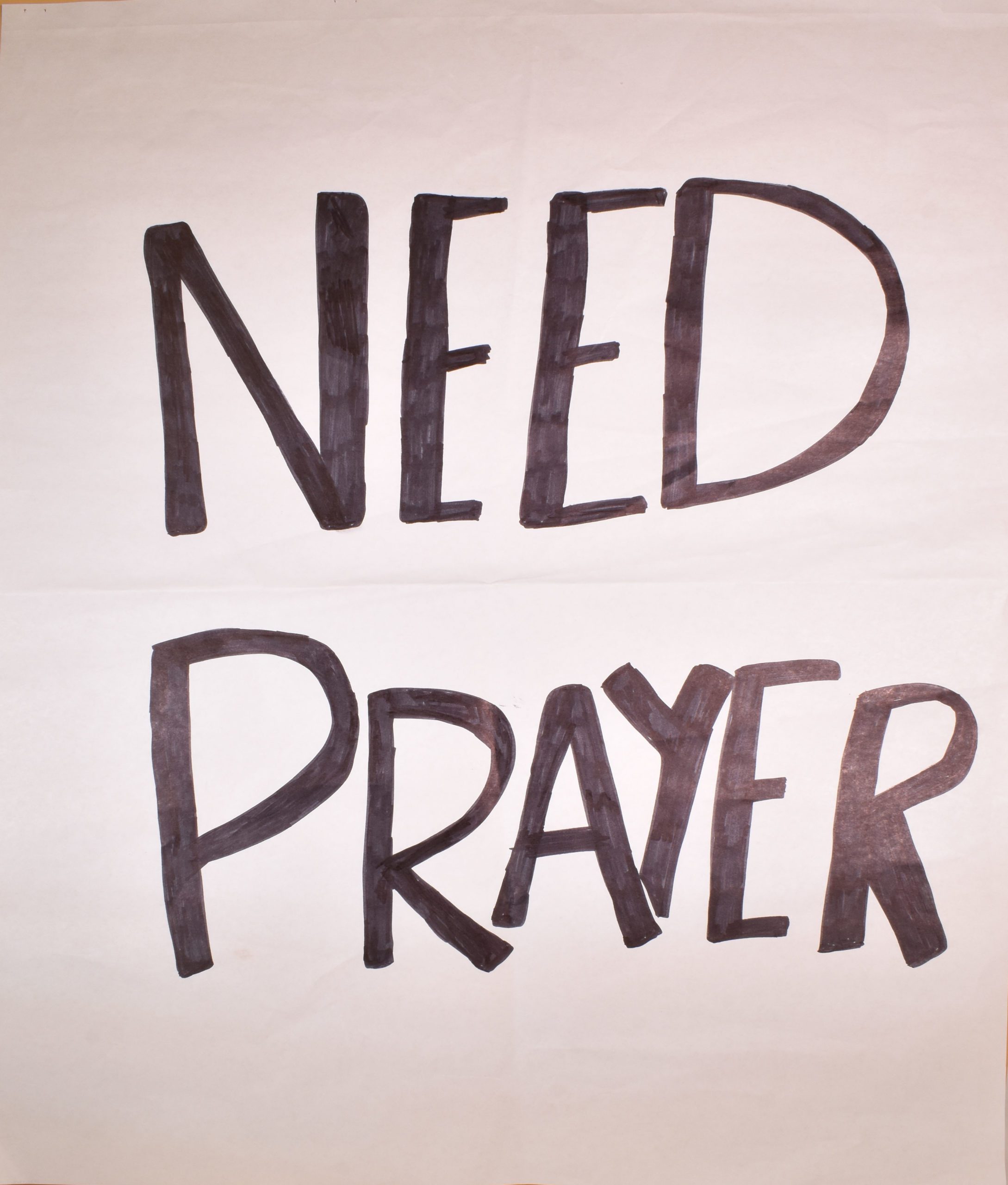 A hand made sign on white paper with "Need Prayer" written in black marker