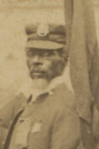 An African American man stands next to a flag on a flagpole. He has a white goatee and wears a cap and jacket with Salvation Army pins and insignia