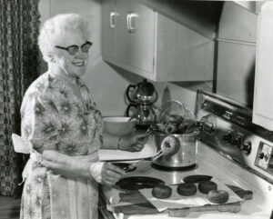 An older woman with white hair and dark glasses smiles and fries doughnuts at her stove.